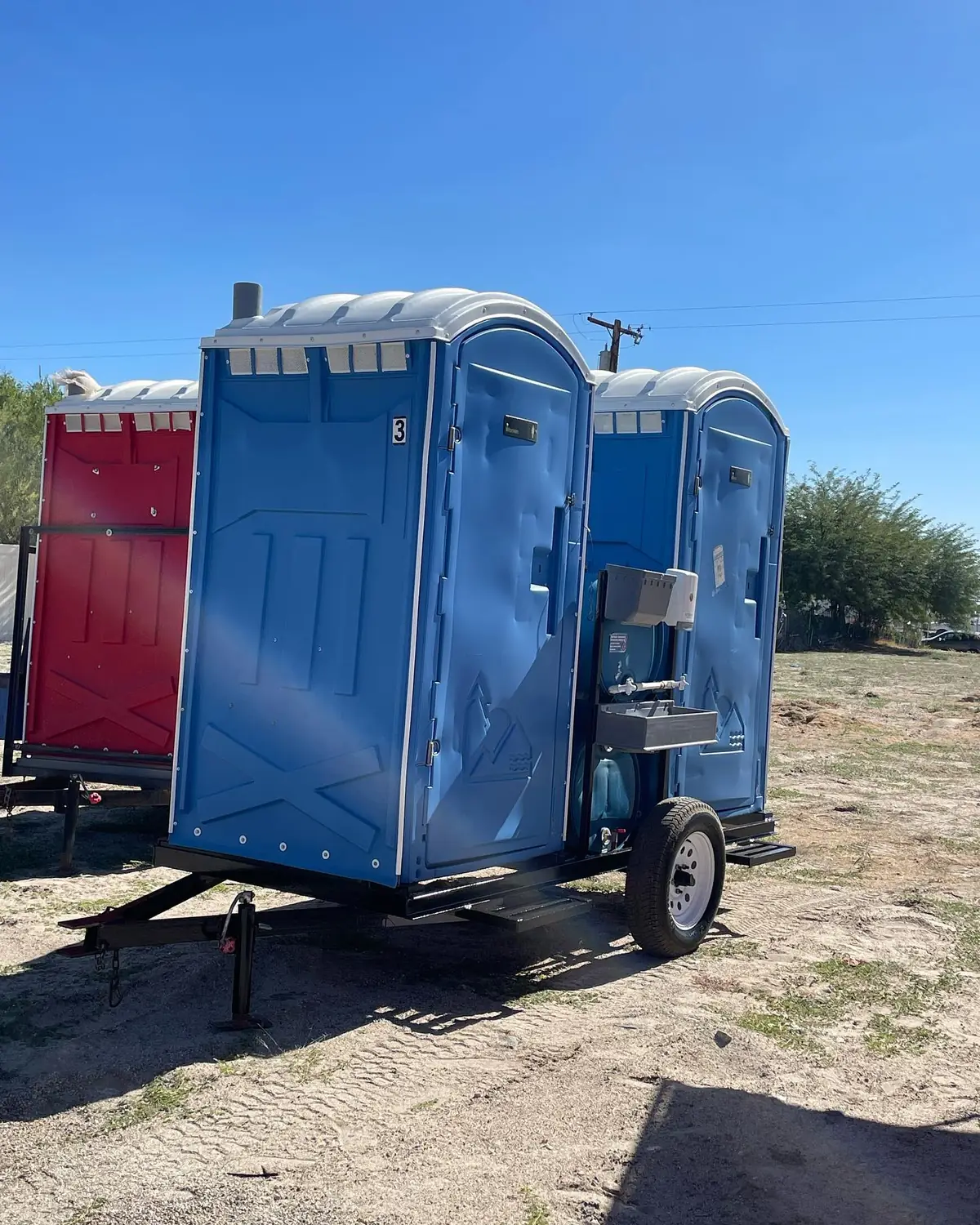 Blue trailer-mounted porta potty with an outside hand-wash sink on a dirt lot